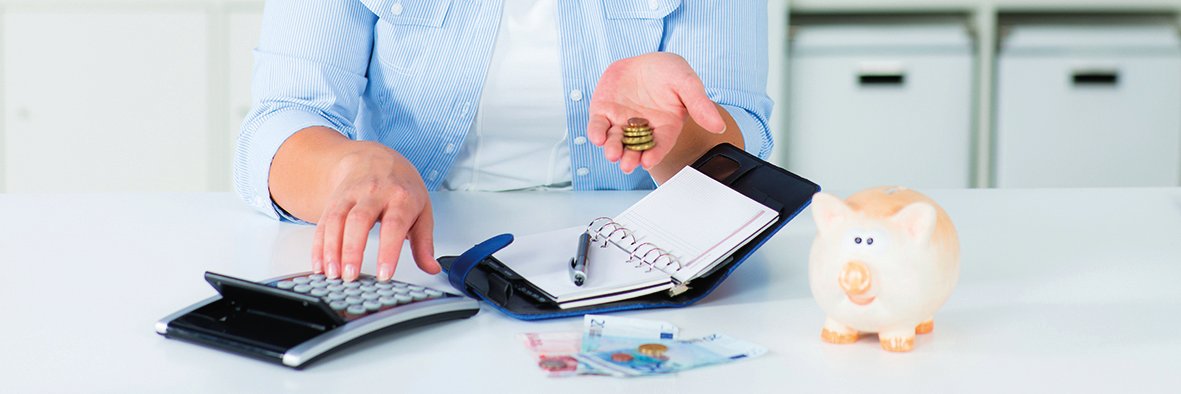 Eine Frau hält Geld in der linken Hand und bedient mit der rechten Hand einen Taschenrechner. Vor Ihr liegt ein aufgeschlagener Organizer und ein Stift. Auf dem Tisch liegen mehrere Geldscheine. Rechts steht ein Sparschwein.