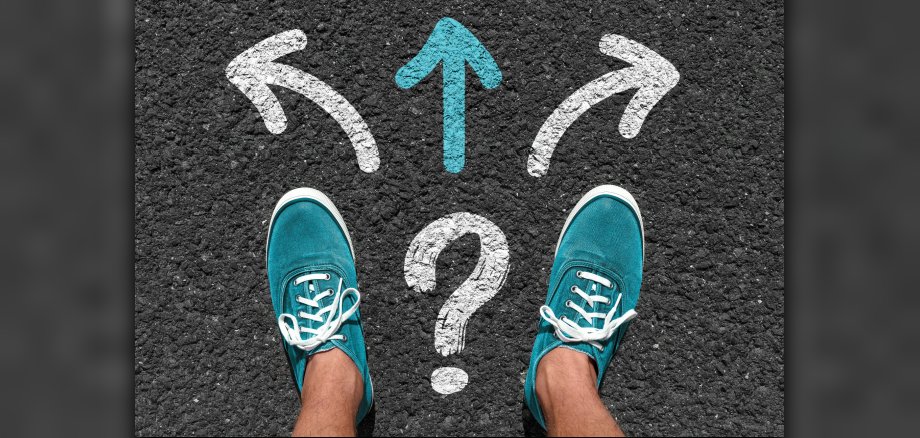 Zwei Männerfüße in türkisen Sneakern auf Asphalt. Vor ihm Richtungspfeile mit Fragezeichen.