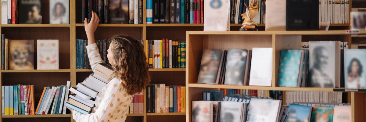 Teenage girl in the middle of a pile of books. A young girl holds books in her hands, bookshelves can be seen in the background. She is surrounded by stacks of books.