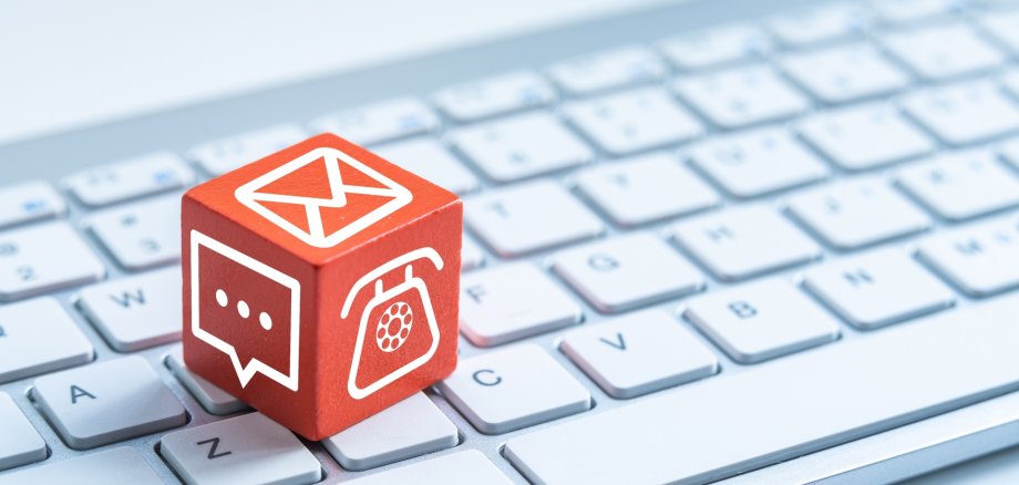 Roter Würfel mit verschiedenen Kontaktsymbolen (Telefon, Chat, Briefumschlag) liegt auf weißer Computertastatur.