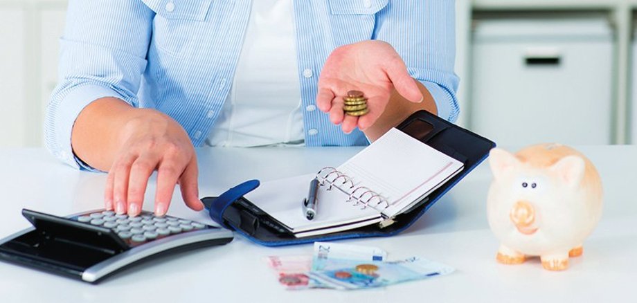 Eine Frau hält Geld in der linken Hand und bedient mit der rechten Hand einen Taschenrechner. Vor Ihr liegt ein aufgeschlagener Organizer und ein Stift. Auf dem Tisch liegen mehrere Geldscheine. Rechts steht ein Sparschwein.