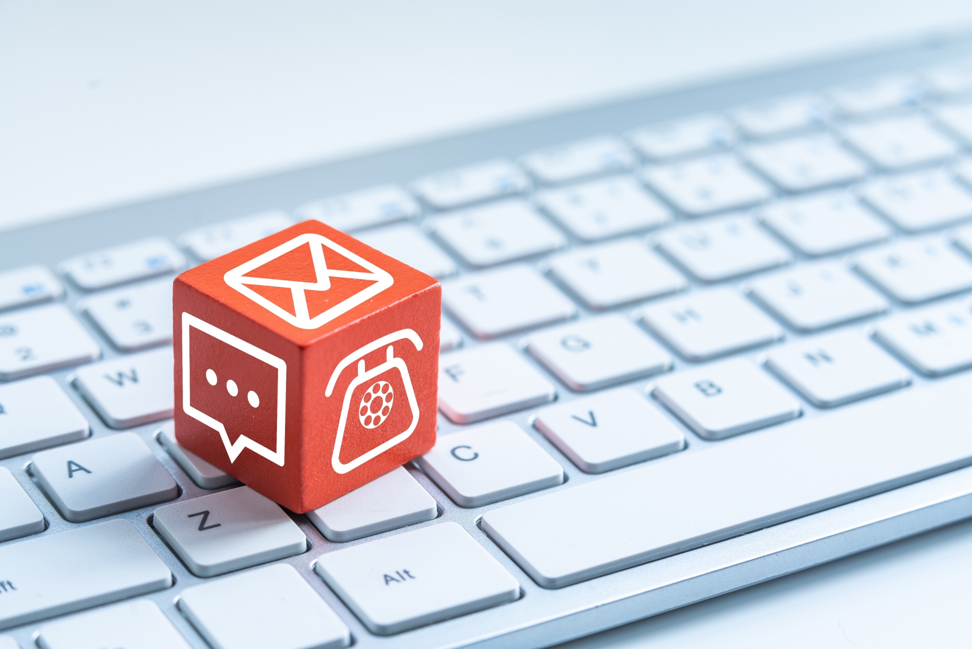 Roter Würfel mit verschiedenen Kontaktsymbolen (Telefon, Chat, Briefumschlag) liegt auf weißer Computertastatur.