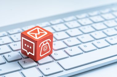 Un cube rouge avec différentes icônes de contact (téléphone, chat, enveloppe) est posé sur un clavier d'ordinateur blanc.