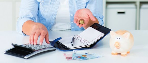 Eine Frau hält Geld in der linken Hand und bedient mit der rechten Hand einen Taschenrechner. Vor Ihr liegt ein aufgeschlagener Organizer und ein Stift. Auf dem Tisch liegen mehrere Geldscheine. Rechts steht ein Sparschwein.