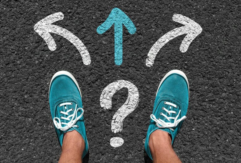 Zwei Männerfüße in türkisen Sneakern auf Asphalt. Vor ihm Richtungspfeile mit Fragezeichen.