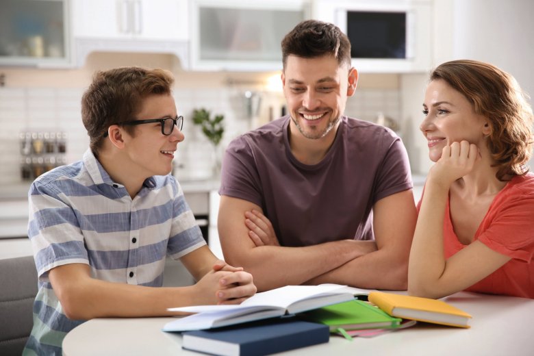Junge sitzt gemeinsam mit seinen Eltern am Tisch. Auf dem Tisch liegen Schulbücher.