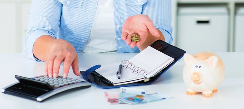 Eine Frau hält Geld in der linken Hand und bedient mit der rechten Hand einen Taschenrechner. Vor Ihr liegt ein aufgeschlagener Organizer und ein Stift. Auf dem Tisch liegen mehrere Geldscheine. Rechts steht ein Sparschwein.